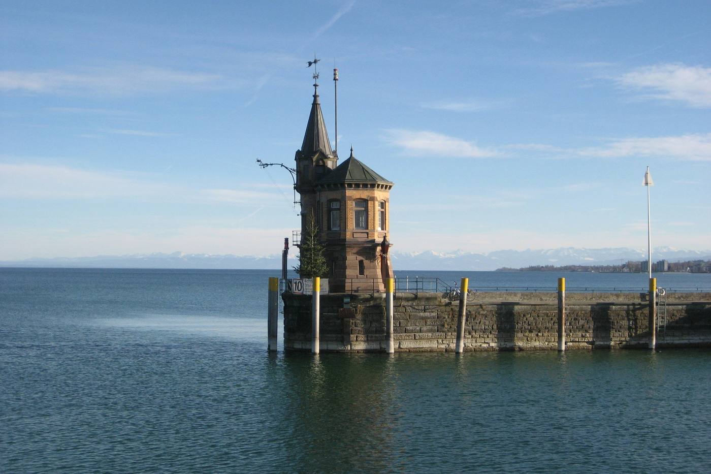 A small brick lighthouse with a pointed roof at the end of a stone pier, the lake and snowy Alps behind